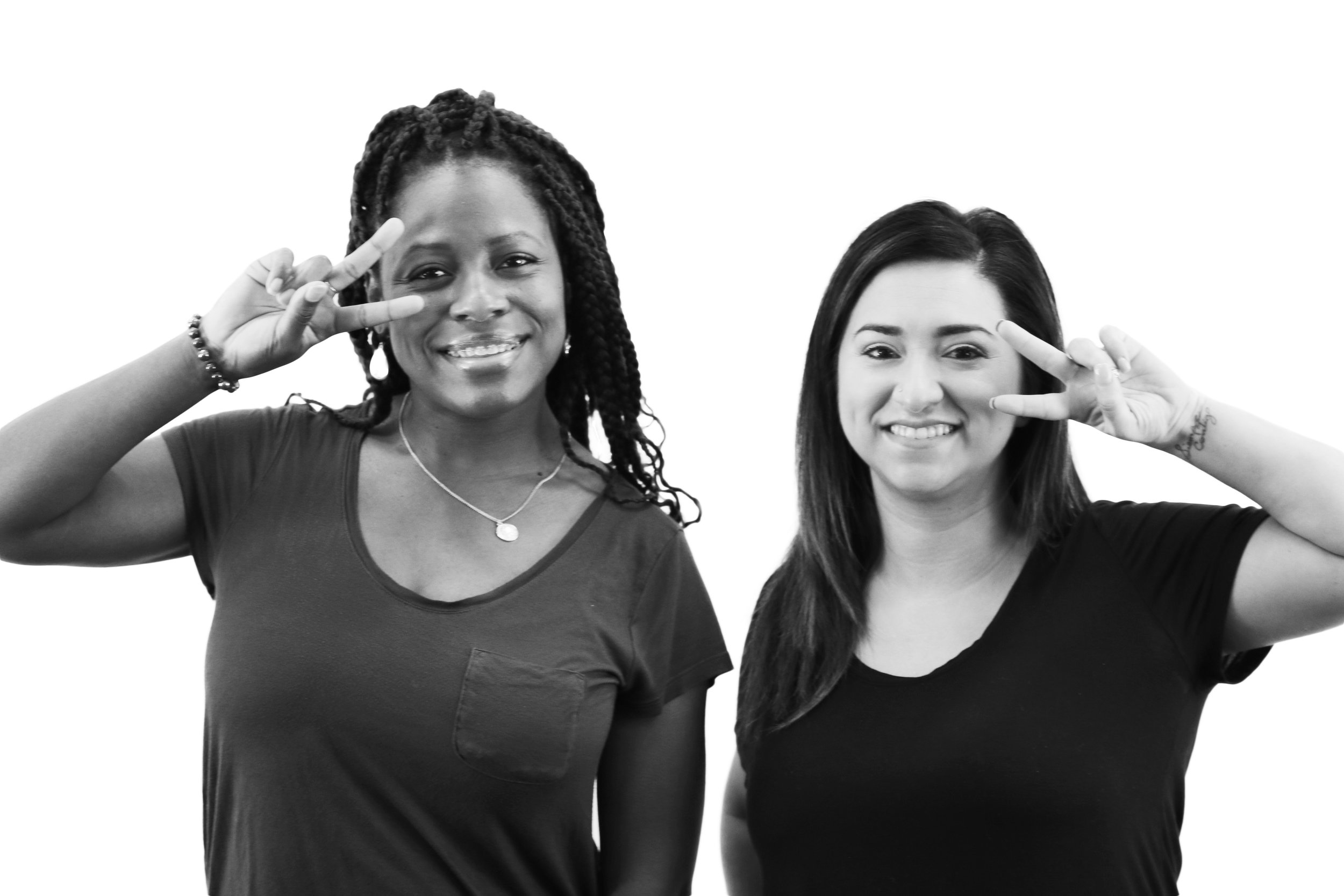 Two women smiling next to each other, putting up peace signs with their hands.