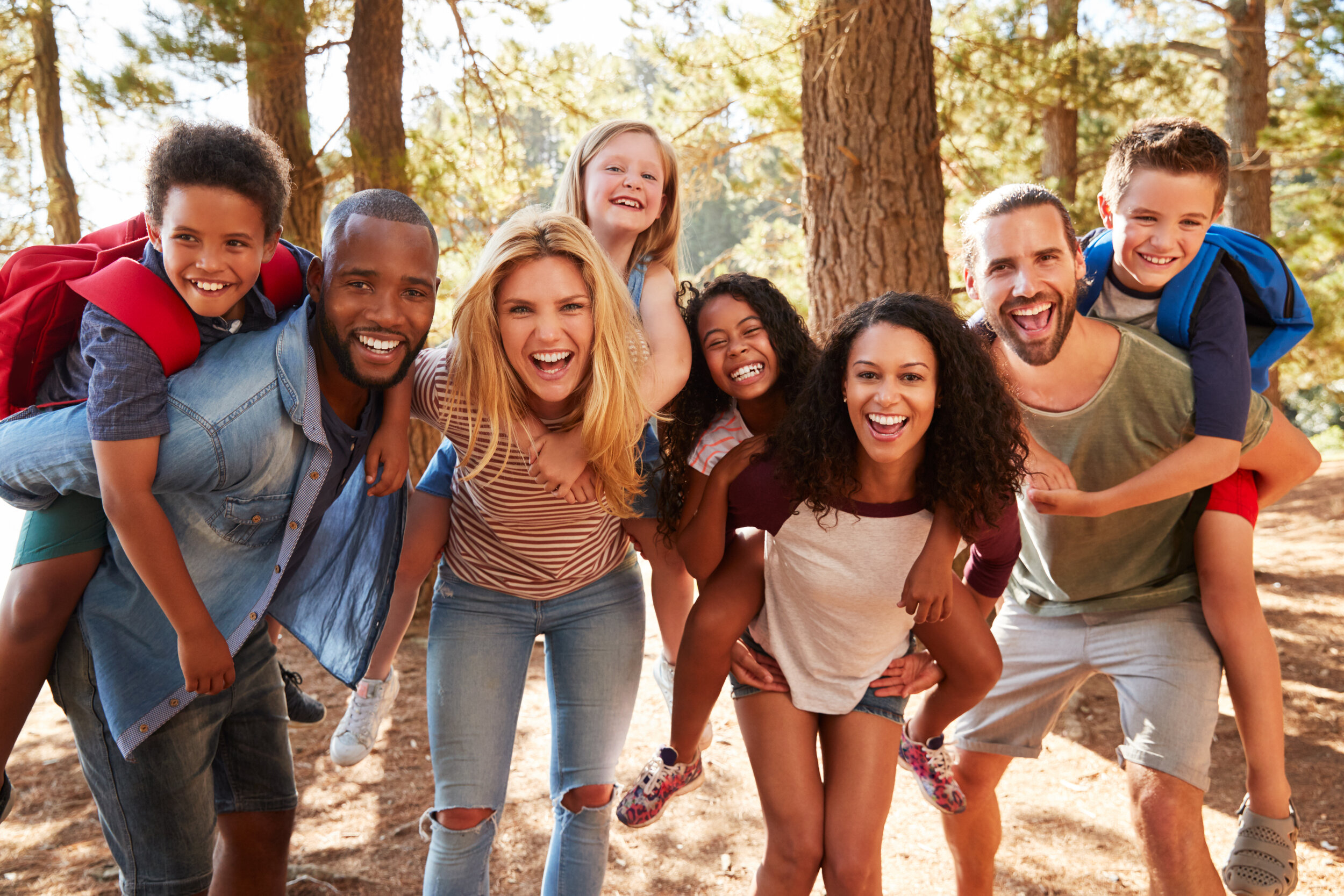 Eight people standing together in the woods with a sunny background.