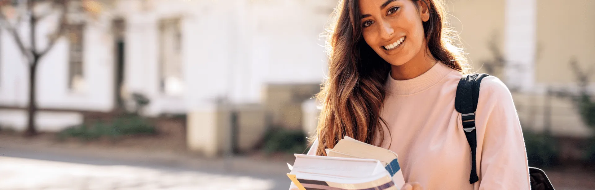 A woman wearing a back pack and holding text books while smiling.